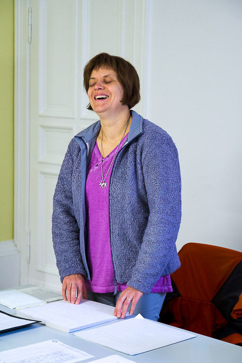 Foto: Kerstin Gaedicke, hinter einem Tisch stehend, hat beide Hände auf ihrem Blindenschriftskript. Sie trägt eine hellgraue Hose, ein fliederfarbenes T-Shirt und darüber eine offene lila Strickjacke. Sie lacht herzlich.